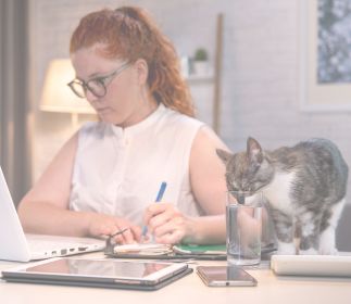 Tipps für das Arbeiten mit Tieren im Homeoffice Eine Frau mit roten Haaren sitzt am Schreibtisch und arbeitet am Laptop. Neben ihr steht ein Glas Wasser, und eine Katze schnuppert daran.