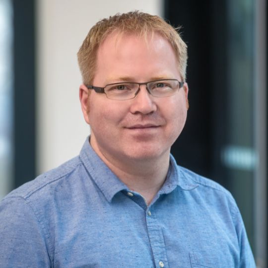 Porträt eines Mannes mit blauer Brille und hellblauem Hemd vor unscharfem Hintergrund