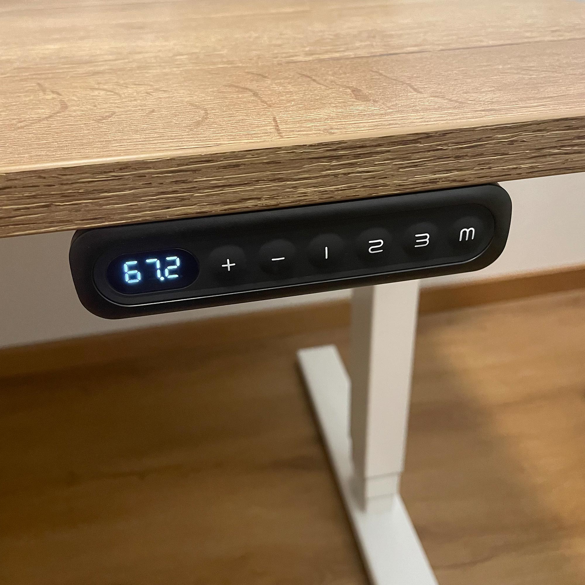 Eine Nahaufnahme eines elektrisch höhenverstellbaren Schreibtisches mit einer Digitalanzeige, die 67,2 anzeigt, und mehreren Tasten unter der hölzernen Tischplatte. Der Schreibtisch hat einen weißen Rahmen und steht auf einem Holzboden.