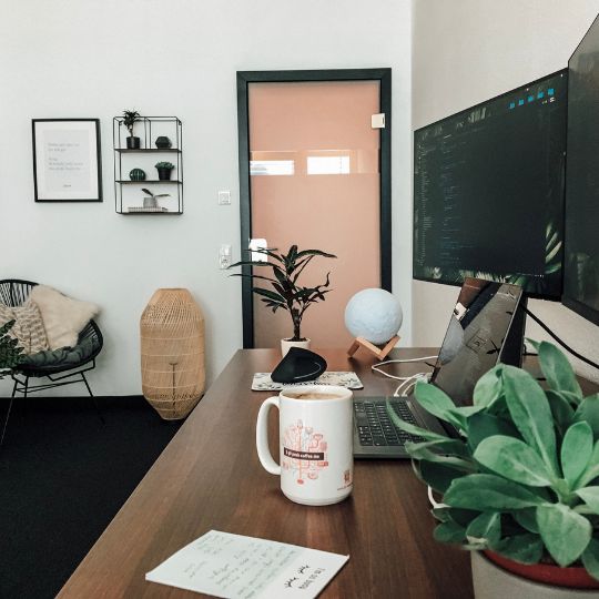 Ein modernes Büro mit einem Schreibtisch, Laptop, Kaffeetasse, Pflanzen und einem Globus. Im Hintergrund sind eine gemütliche Sitzecke und eine Tür zu sehen.