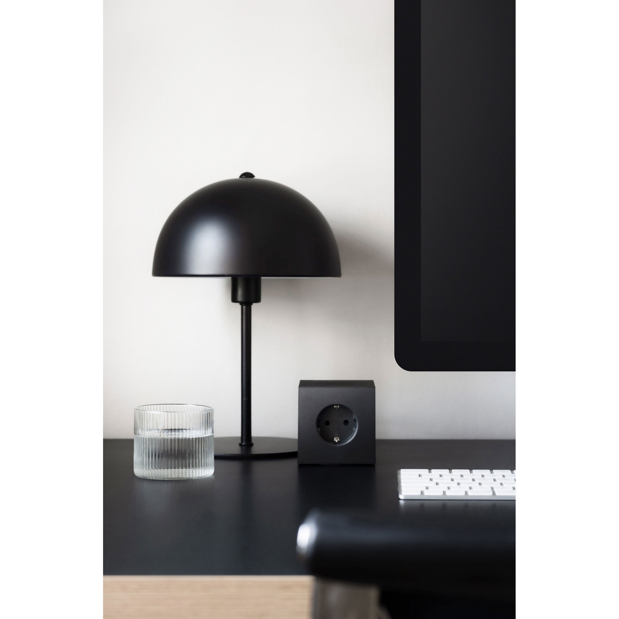 Schreibtisch-Szene mit AVOLT Square 1 Steckdosenwürfel in Schwarz neben Lampe, Glas und Monitor – stilvolles, modernes Arbeitsplatz-Setup