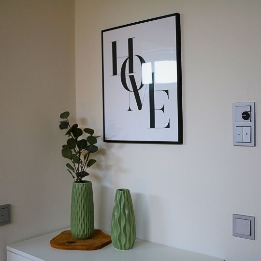 Wanddekoration mit HOME-Schriftzug und zwei grünen Vasen auf Sideboard.