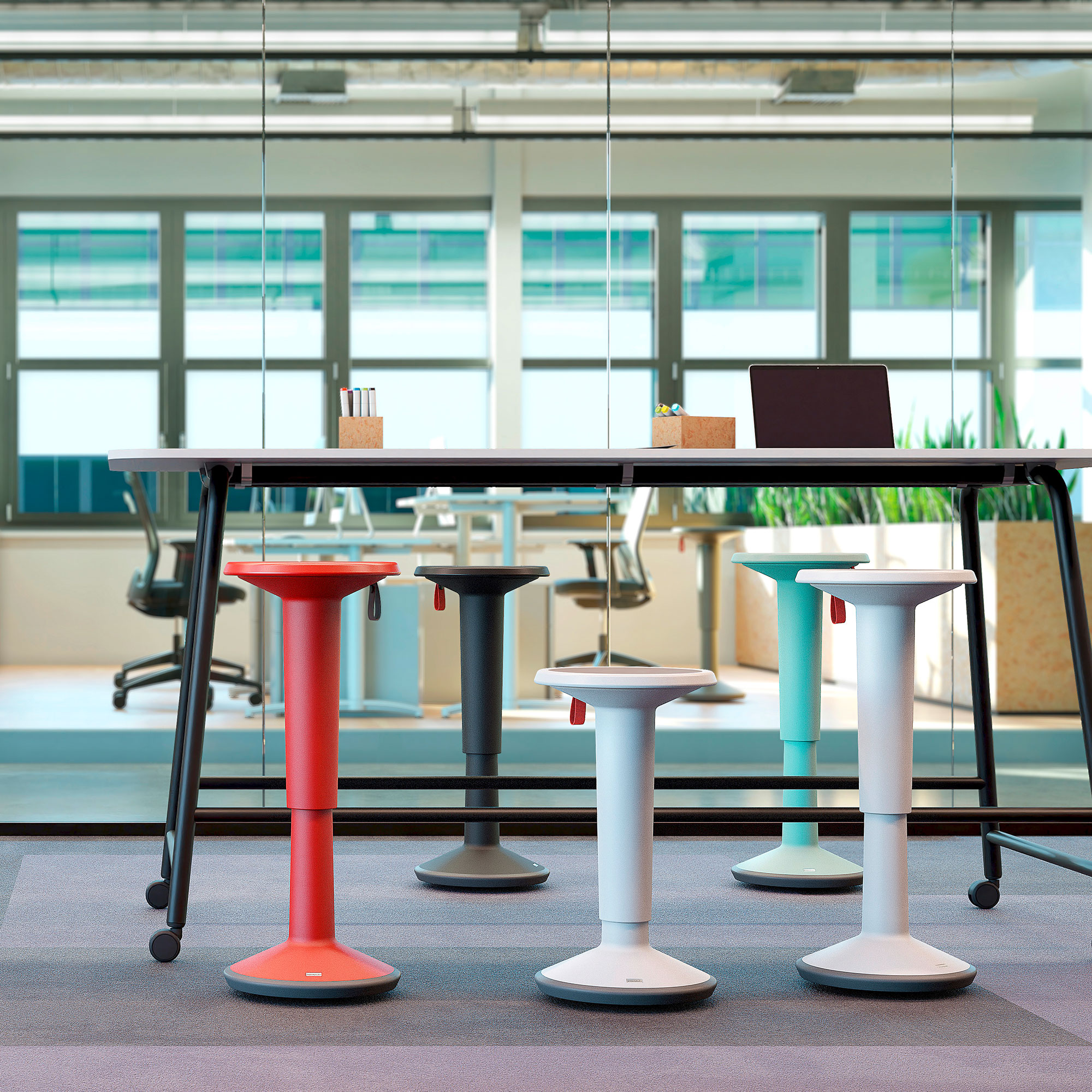 Steh-Sitzhocker UP Ein moderner Büroraum mit einem langen Tisch und vier bunten Stehhilfen in Rot, Schwarz, Weiß und Blau. Im Hintergrund sind Bürostühle und Pflanzen zu sehen.