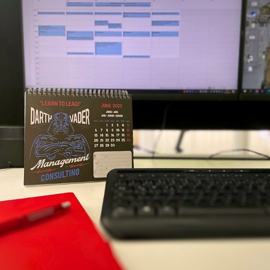Ein Tisch mit einem Kalender für Juni 2022, der Darth Vader zeigt, und einem Computer im Hintergrund mit einem Kalender. Zudem sind ein rotes Notizbuch und ein Drehstift sichtbar.