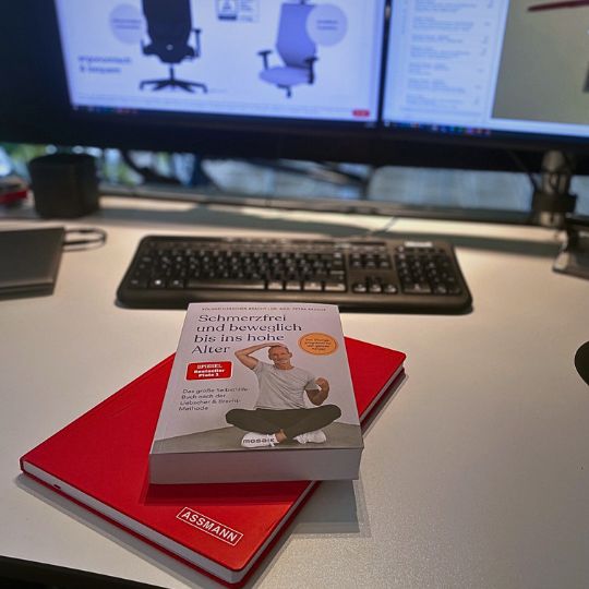 Buch und rotes Notizbuch auf einem Schreibtisch vor zwei Monitoren.