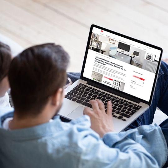 Eine Person sitzt auf einem Sofa und nutzt einen Laptop, auf dem eine Webseite mit Möbeln angezeigt wird.