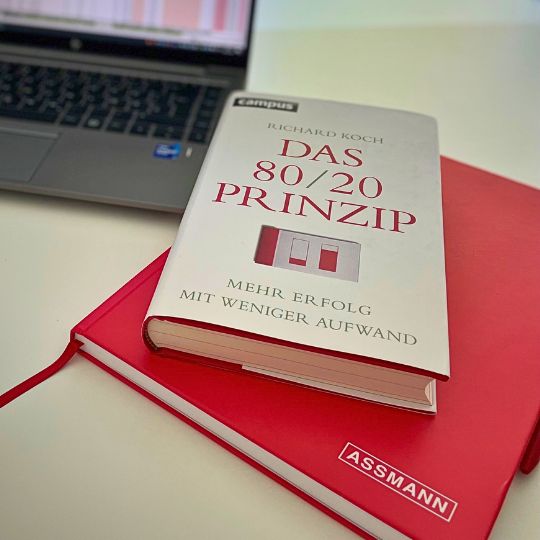 Das Buch 'Das 80/20 Prinzip' liegt auf einem roten ASSMANN-Notizbuch neben einem geöffneten Laptop