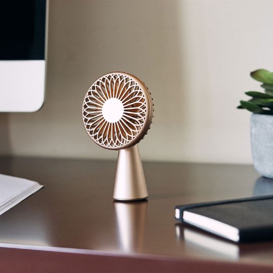 Kleiner goldfarbener Tischventilator auf einem braunen Schreibtisch neben Monitor und Notizbuch