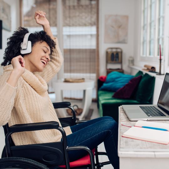 Eine fröhliche Person mit Kopfhörern sitzt in einem Rollstuhl und genießt Musik am Laptop in einem hellen Raum.