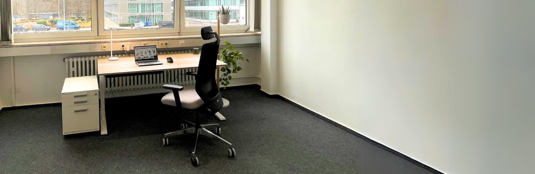 Großzügiger Büroraum mit höhenverstellbarem Schreibtisch, schwarzem Drehstuhl und mobilem Rollcontainer.
