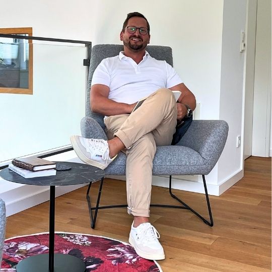 Ein Mann sitzt in einem modernen Sessel, trägt ein weißes Hemd und beige Hosen. Neben ihm steht ein kleiner Tisch mit einem Notizbuch. Der Raum hat Holzboden und ist hell.