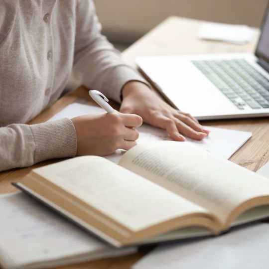 Person schreibt Notizen in ein Buch neben einem Laptop auf einem Holztisch.