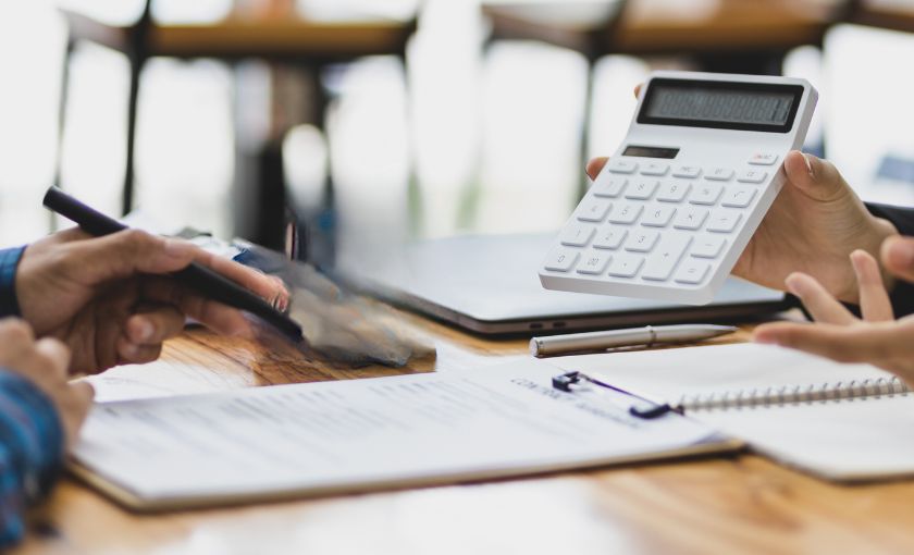 Person hält einen weißen Taschenrechner über einem Tisch mit Laptop und Unterlagen – Gespräch über Finanzen oder Planungsszenario