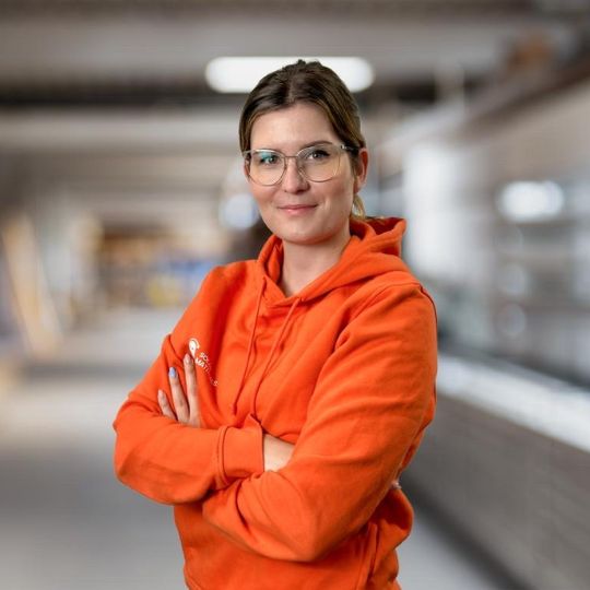 Eine junge Frau mit Brille trägt einen orangefarbenen Kapuzenpullover und steht mit verschränkten Armen in einer hellen Umgebung.