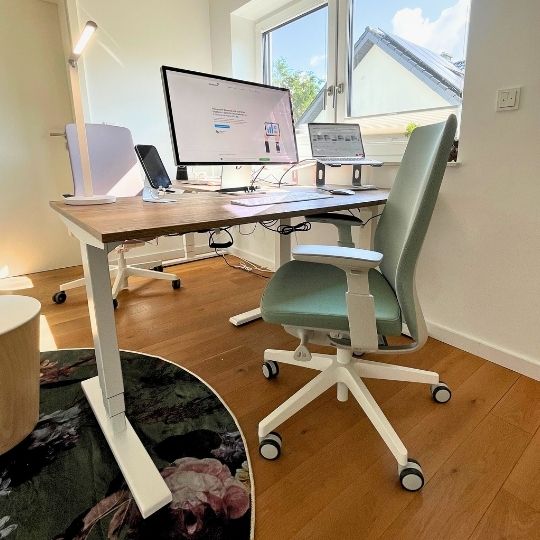 Höhenverstellbarer Schreibtisch mit Holzplatte und Monitoren, daneben ein grüner Bürostuhl.