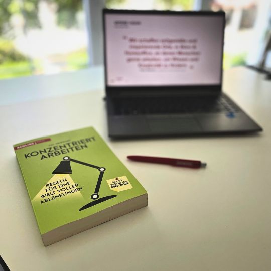Ein Buch mit dem Titel 'KONZENTRIERT ARBEITEN' liegt auf einem Tisch, neben einem Laptop, auf dessen Bildschirm ein Zitat zu sehen ist. Ein roter Stift liegt ebenfalls auf dem Tisch.