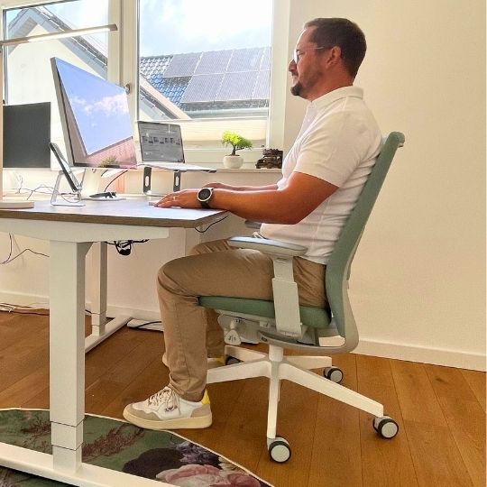 Ein Mann sitzt an einem Schreibtisch mit mehreren Monitoren und arbeitet. Er trägt ein weißes Poloshirt und beige Hosen. In der Umgebung sind ein Fenster und einige Pflanzen sichtbar.