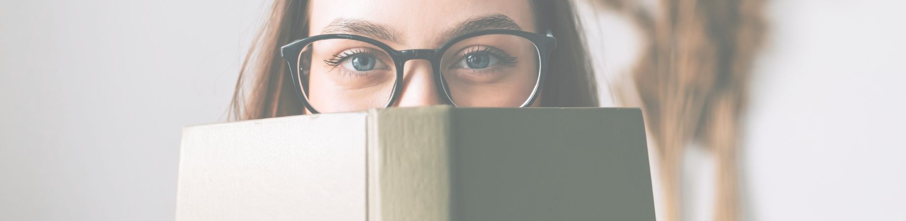 Frau mit Brille blickt über ein geöffnetes Buch, Hintergrund unscharf.
