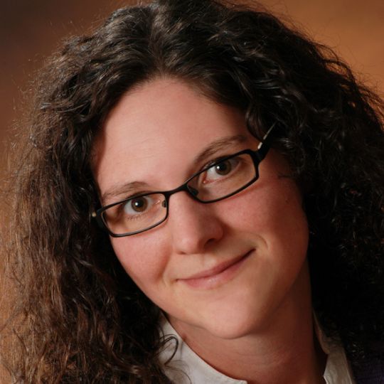 Ein Porträt einer Person mit lockigem, braunem Haar und Brille, die lächelt.