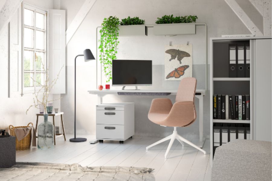 Ein modernes Arbeitszimmer mit einem Schreibtisch, einem Computerbildschirm, einer rosa Stuhls, einem Rollcontainer, einer Stehlampe und Pflanzen.