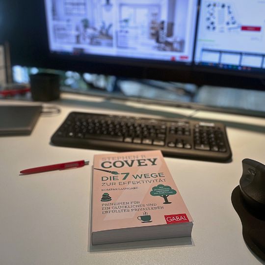 Ein Schreibtisch mit dem Buch 'Die 7 Wege zur Effektivität' von Stephen Covey, einem roten Stift und einem Computerbildschirm im Hintergrund.