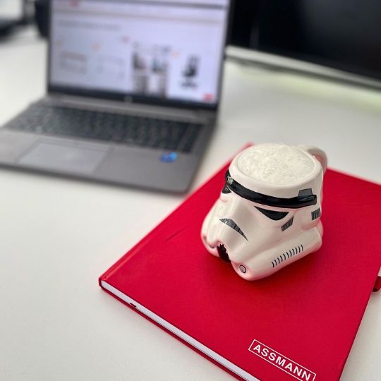 Stormtrooper-Tasse auf rotem ASSMANN-Notizbuch vor geöffnetem Laptop auf weißem Schreibtisch
