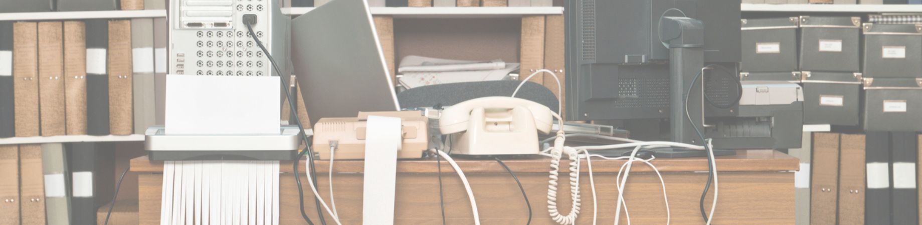 Unordentlicher Schreibtisch mit Telefon, Aktenordnern und vielen Kabeln vor einem Regal.