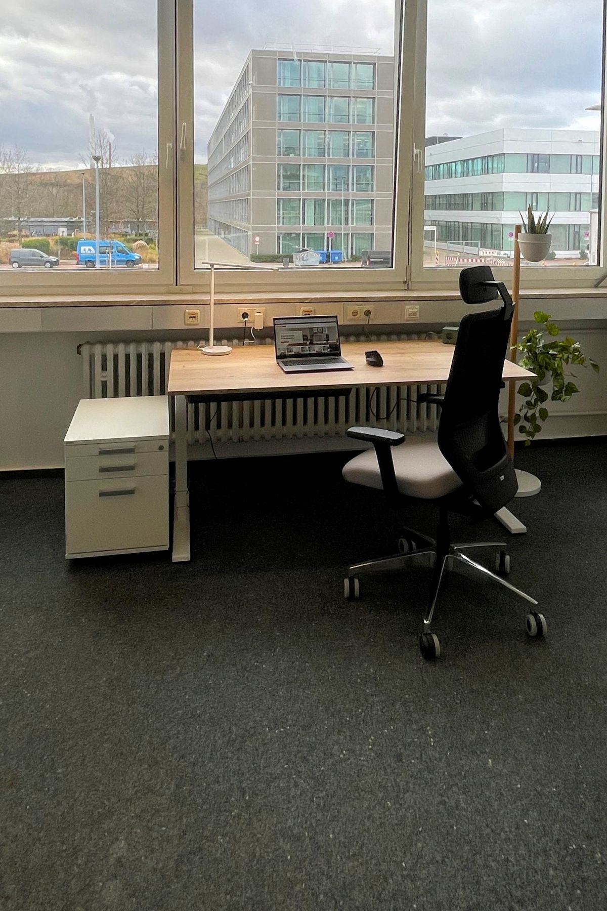 Ein Arbeitsplatz mit einem Schreibtisch, einem Computer, einem Bürostuhl und Fenstern, die den Blick auf ein modernes Gebäude freigeben.