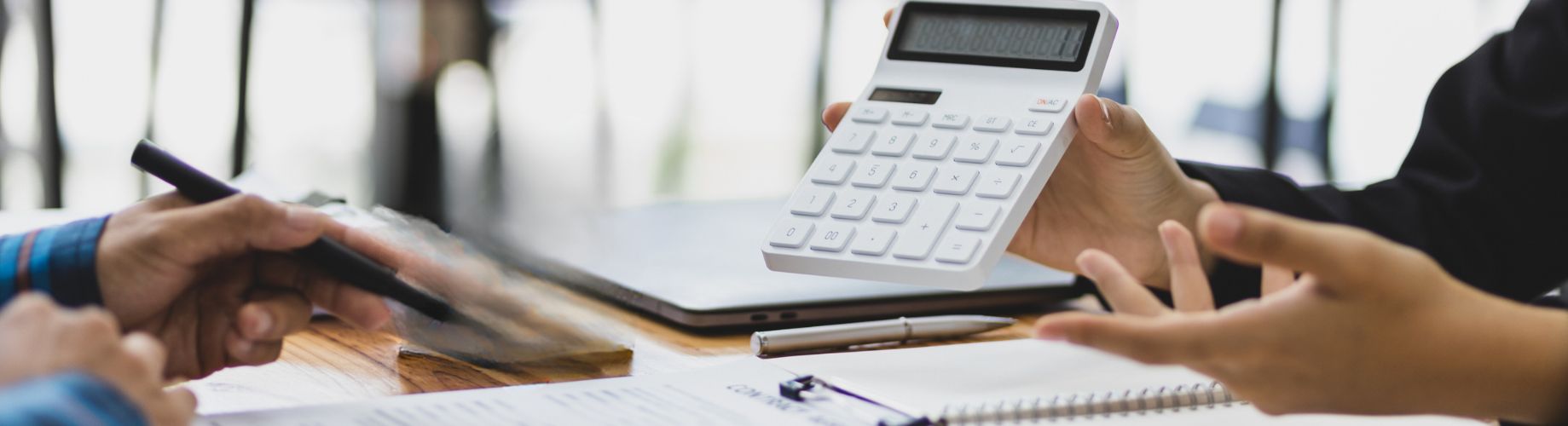 Person hält einen weißen Taschenrechner über einem Tisch mit Laptop und Unterlagen – Gespräch über Finanzen oder Planungsszenario
