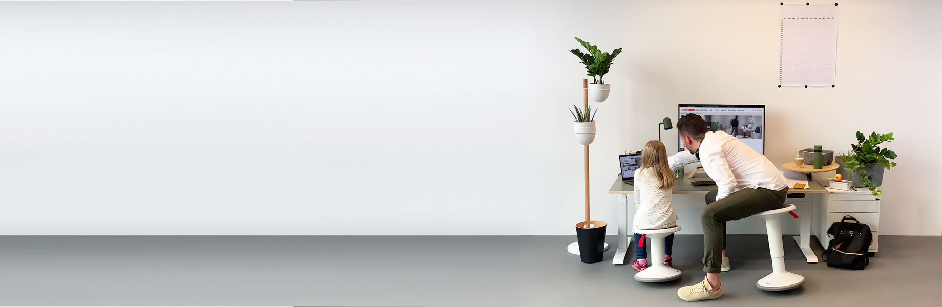 Ein Vater sitzt mit seiner Tochter an einem modernen Schreibtisch und schaut auf einen Computerbildschirm. Um sie herum sind Pflanzen und Büromaterialien.