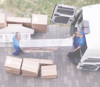 Speditionsversand bei ASSMANN HOME Zwei Personen tragen einen großen, transparenten Gegenstand aus einem Lieferwagen, umgeben von Kartons.