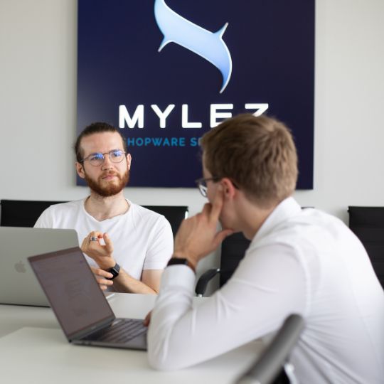 Zwei Mitarbeiter im Gespräch vor Firmenlogo 8mylez mit Laptops in einem modernen Konferenzraum.