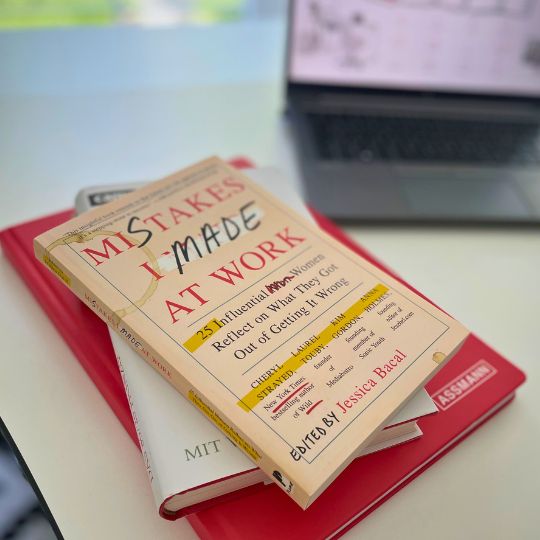 Ein Buch mit dem Titel 'Mistakes I Made at Work' liegt auf einem roten Ordner und einem weiteren Buch, neben einem Laptop.
