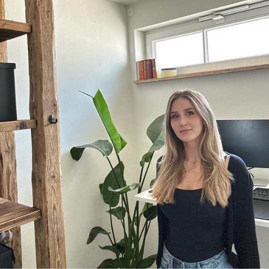Junge Frau steht in modernem Homeoffice mit Pflanze und Holzregal