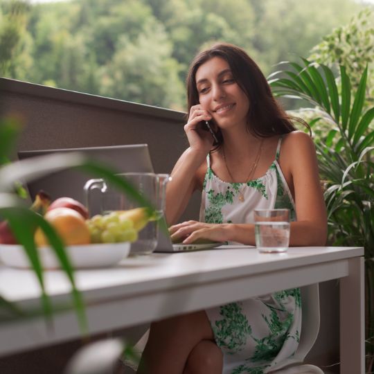 Eine Frau sitzt auf einem Balkon, telefoniert, lächelt und arbeitet am Laptop. Auf dem Tisch stehen ein Glas Wasser, ein Krug und eine Schüssel mit Obst.